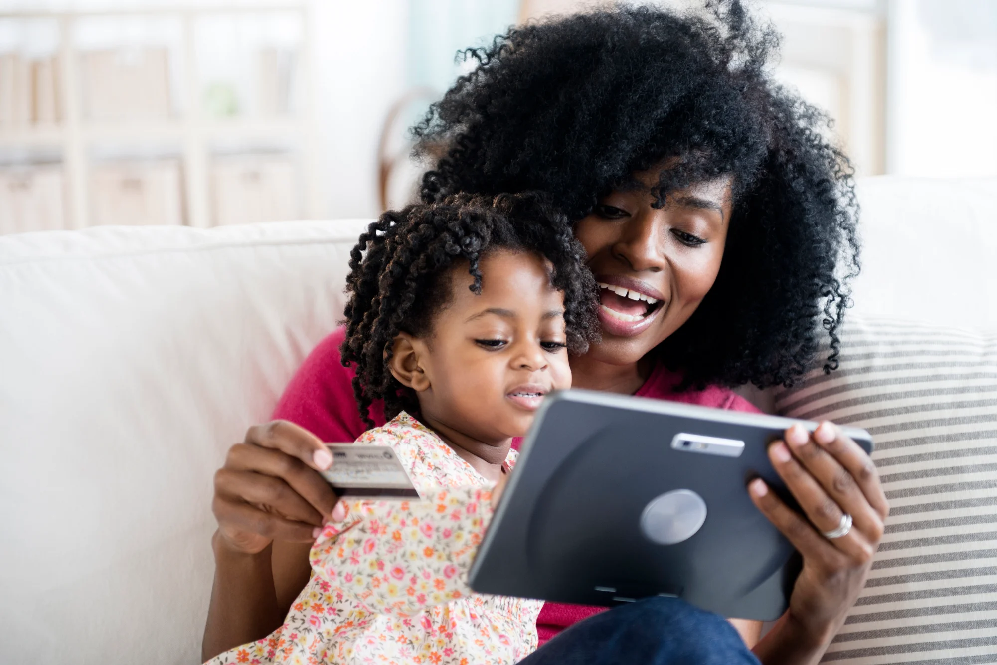 A smiling woman and her young daughter sit together on a sofa, looking at a tablet while holding a credit card. They are empowered and ready to manage their next steps with confidence.