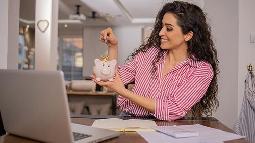 Woman with bills, laptop and calculator in front of her and adding money in a piggy bank.