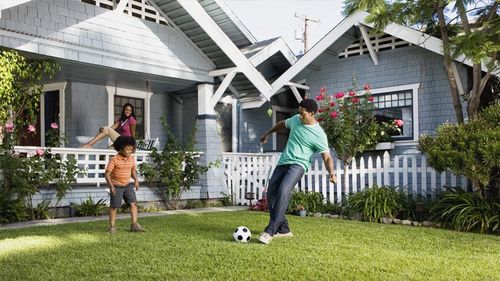 Father and son playing soccer on the front lawn while mom watches from the porch.